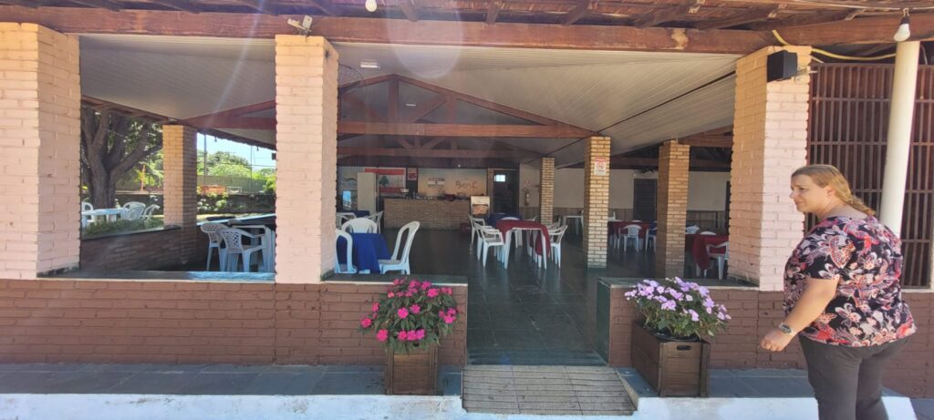 Área coberta do bar e restaurante, com pilares de tijolo, mesas e cadeiras, floreiras e rampa de acesso.