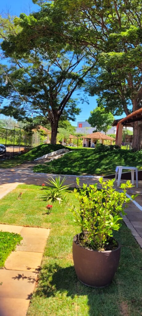 Área verde arborizada, com caminho de pedra, quiosques ao fundo e quadra cercada à esquerda.