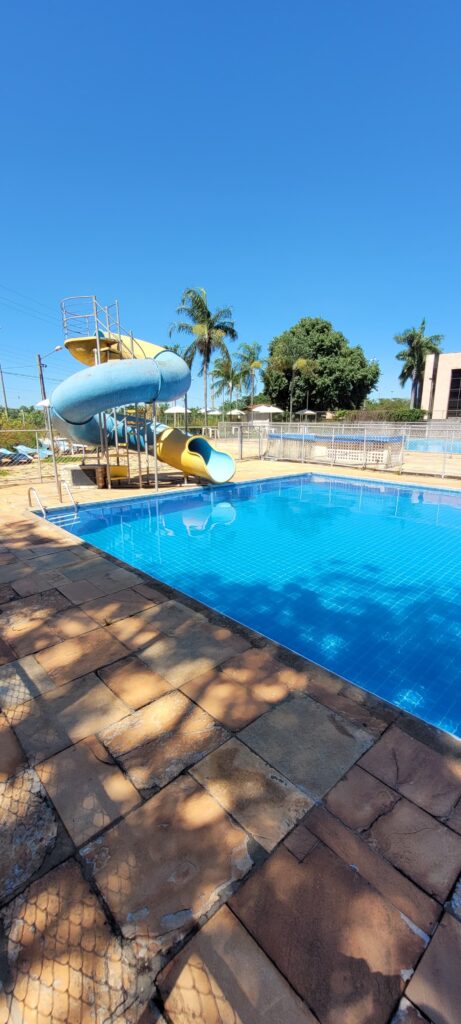 Piscina de adultos com toboágua azul e amarelo, cercada por palmeiras, em dia de sol.